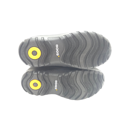 A pair of black BOGS boots shown from the sole view, featuring a textured black rubber tread with a bright yellow circular logo accent and white BOGS branding.
