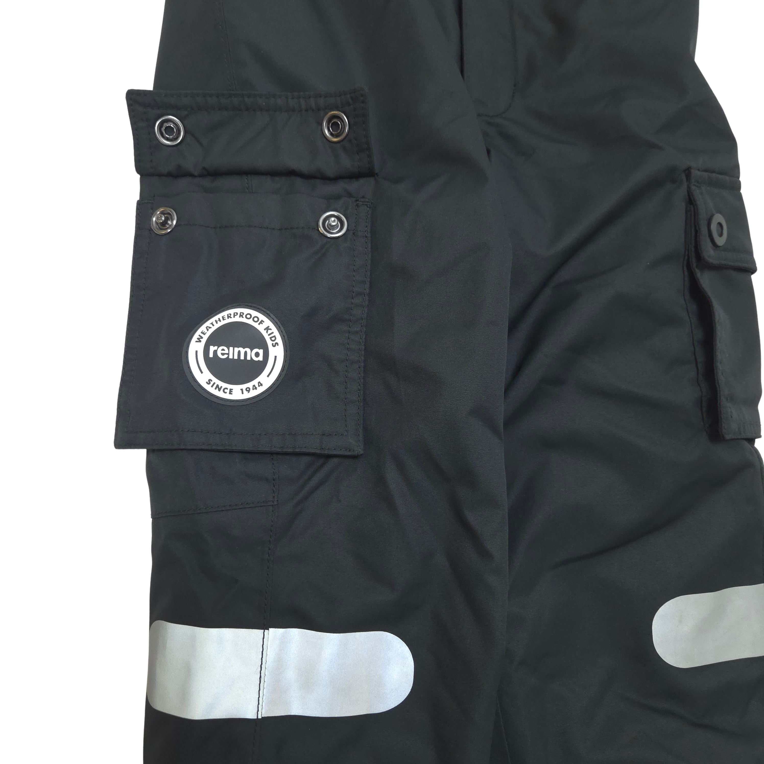Close-up of black shell pants with reflective silver stripes and a white circular Reima logo patch on a cargo pocket.