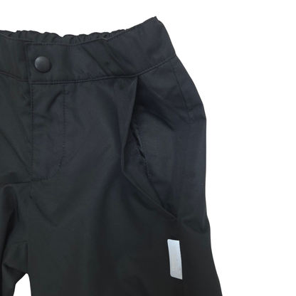 Close-up of black outdoor pants showing the waistband with a snap button, side pocket, and a small white rectangular reflective tag on a plain white background.