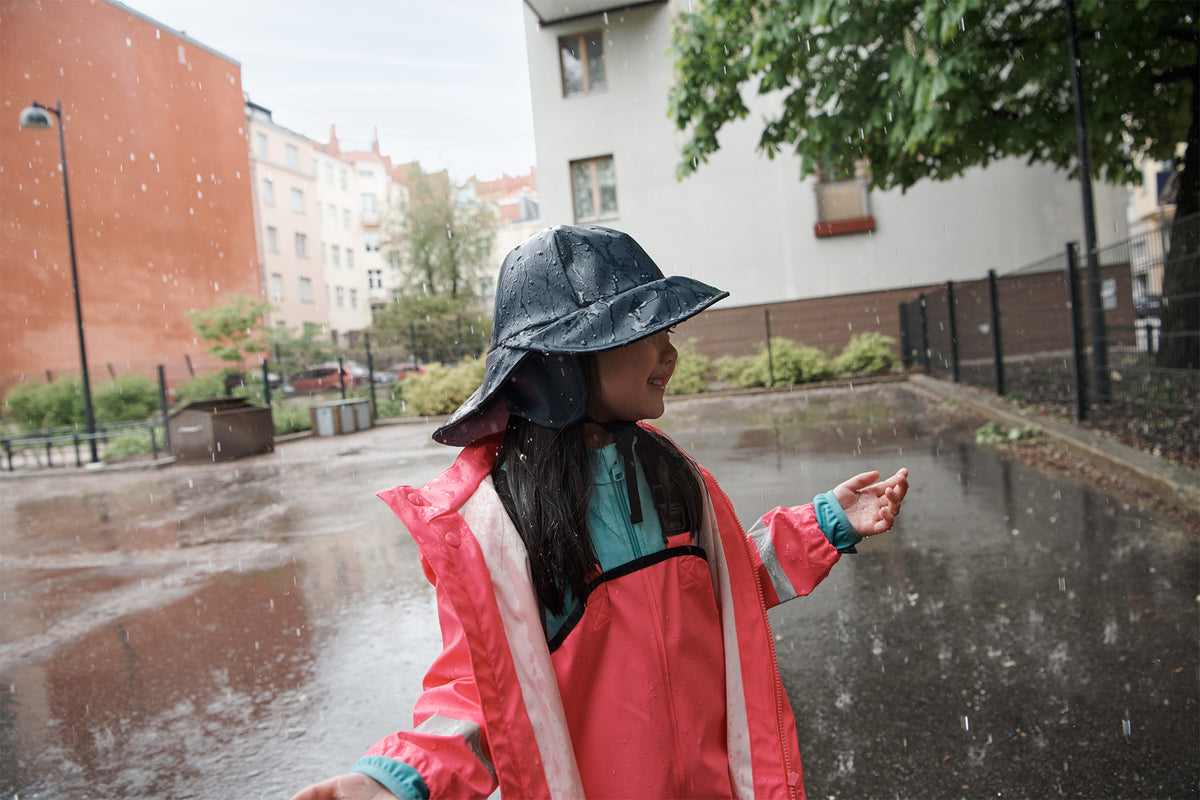 A child wearing a bright pink raincoat and a dark blue rain hat stands in the rain on a wet city street, looking to the side.