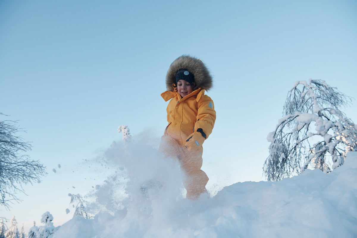 A child wearing a bright yellow winter parka with a fur-trimmed hood and matching snow pants stands in deep snow, kicking up a spray of powder against a clear blue sky.