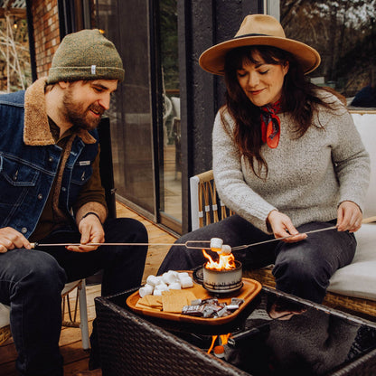 A man and a woman sit by a fire pit on a patio, roasting marshmallows on sticks. The man wears a green beanie and denim jacket with a shearling collar, and the woman wears a tan wide-brim hat, a gray knit sweater, and a red neck scarf.