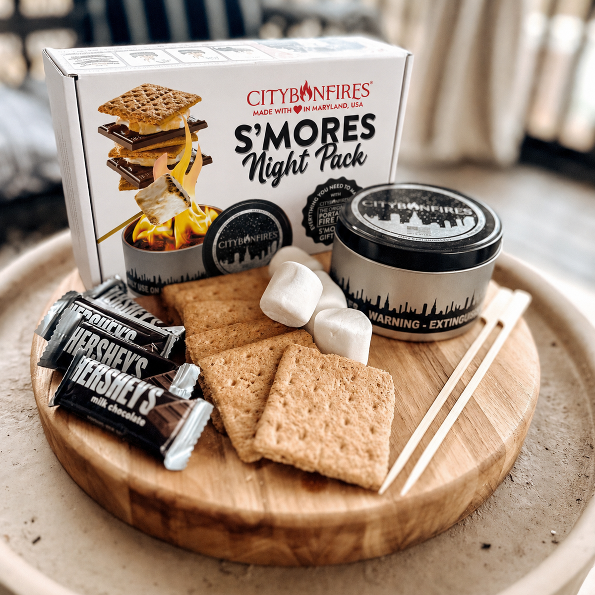 City of Fires S'mores Night Pack gift set arranged on a round wooden tray, including a white branded box, a black-lidded tin, Hershey's chocolate bars, graham crackers, marshmallows, and wooden sticks, photographed in a real indoor setting.