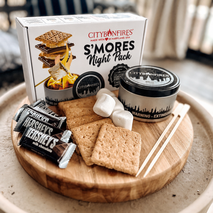 City of Fires S'mores Night Pack gift set arranged on a round wooden tray, including a white branded box, a black-lidded tin, Hershey's chocolate bars, graham crackers, marshmallows, and wooden sticks, photographed in a real indoor setting.