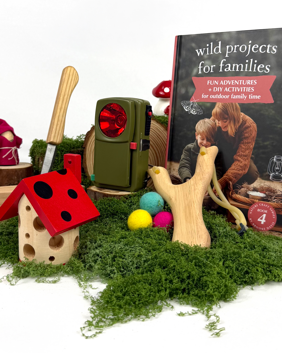 A styled product display featuring a book titled 'wild projects for families' standing upright next to a wooden mushroom house with a red roof, a green lantern, a wooden branch-shaped tool, colorful balls, moss, and a small gnome figure on a white background.