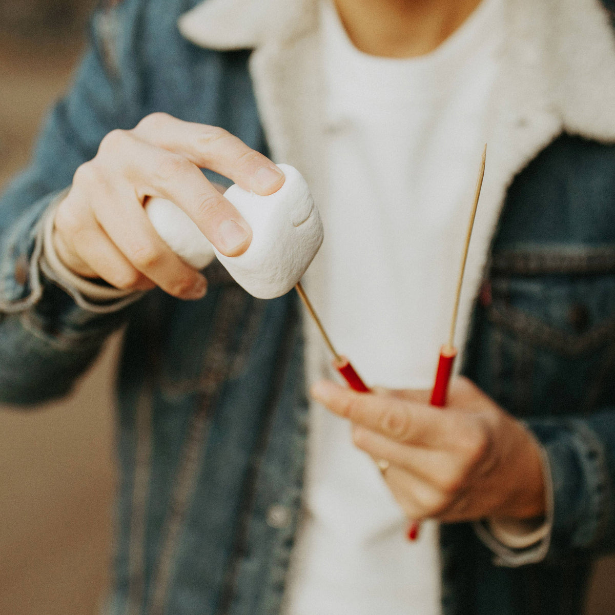 A close-up lifestyle shot of a person roasting a marshmallow on a metal skewer with red handles, wearing a white t-shirt and a blue denim jacket.