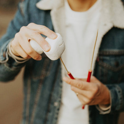 A close-up lifestyle shot of a person roasting a marshmallow on a metal skewer with red handles, wearing a white t-shirt and a blue denim jacket.