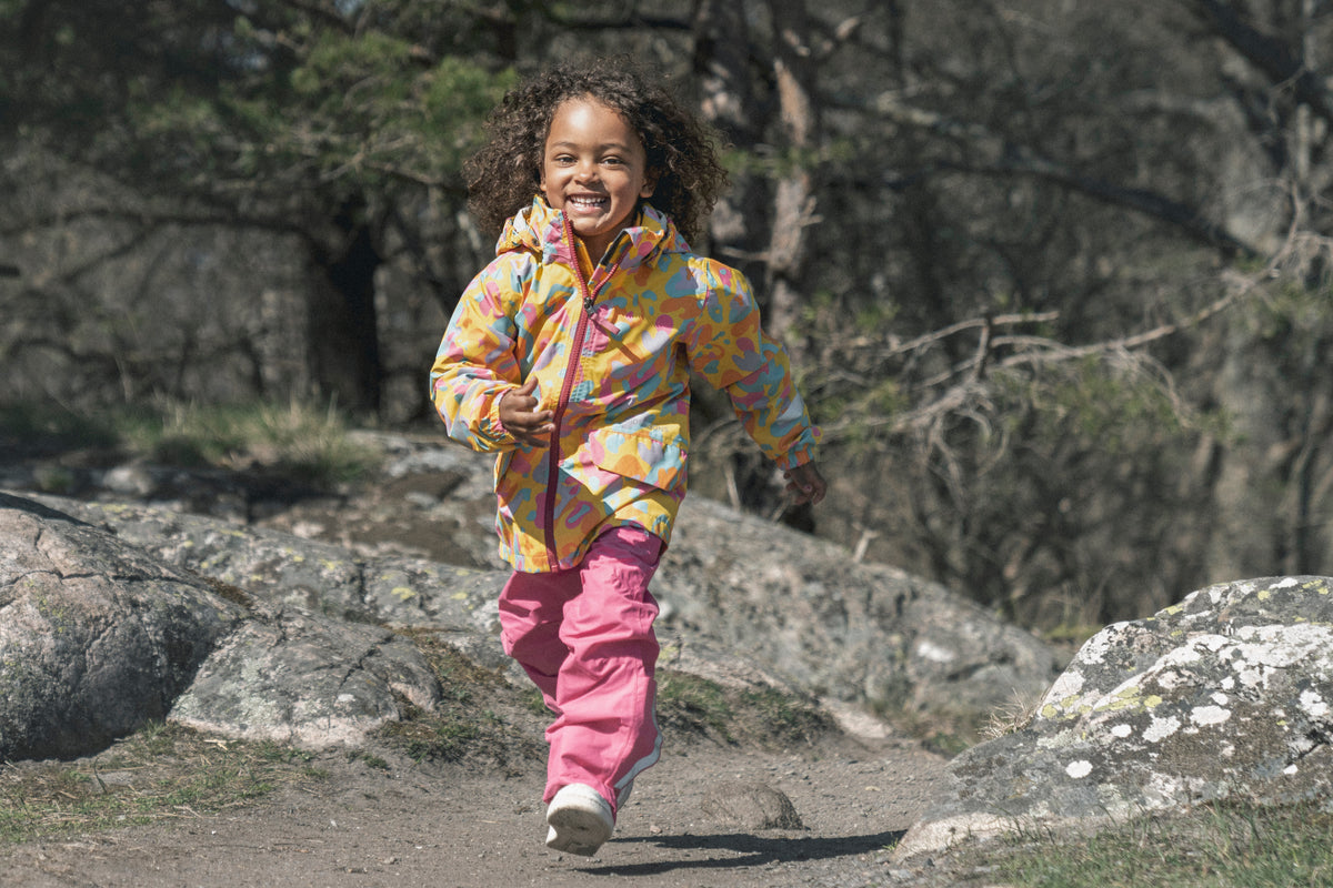Child in colorful jacket and pink pants running on a rocky path with trees in the background
