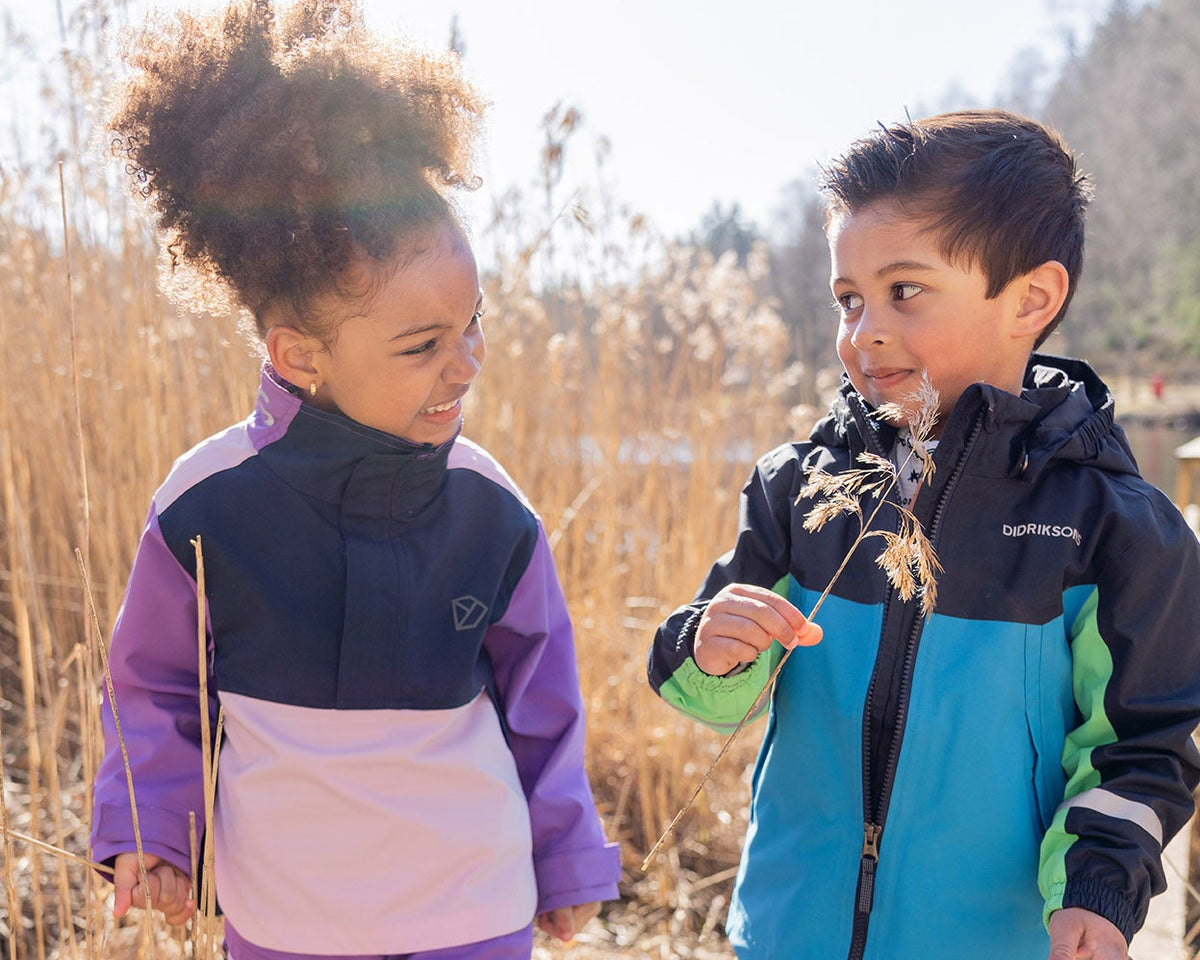 Two children in colorful winter jackets standing on a wooden boardwalk with dry grass and trees in the background.