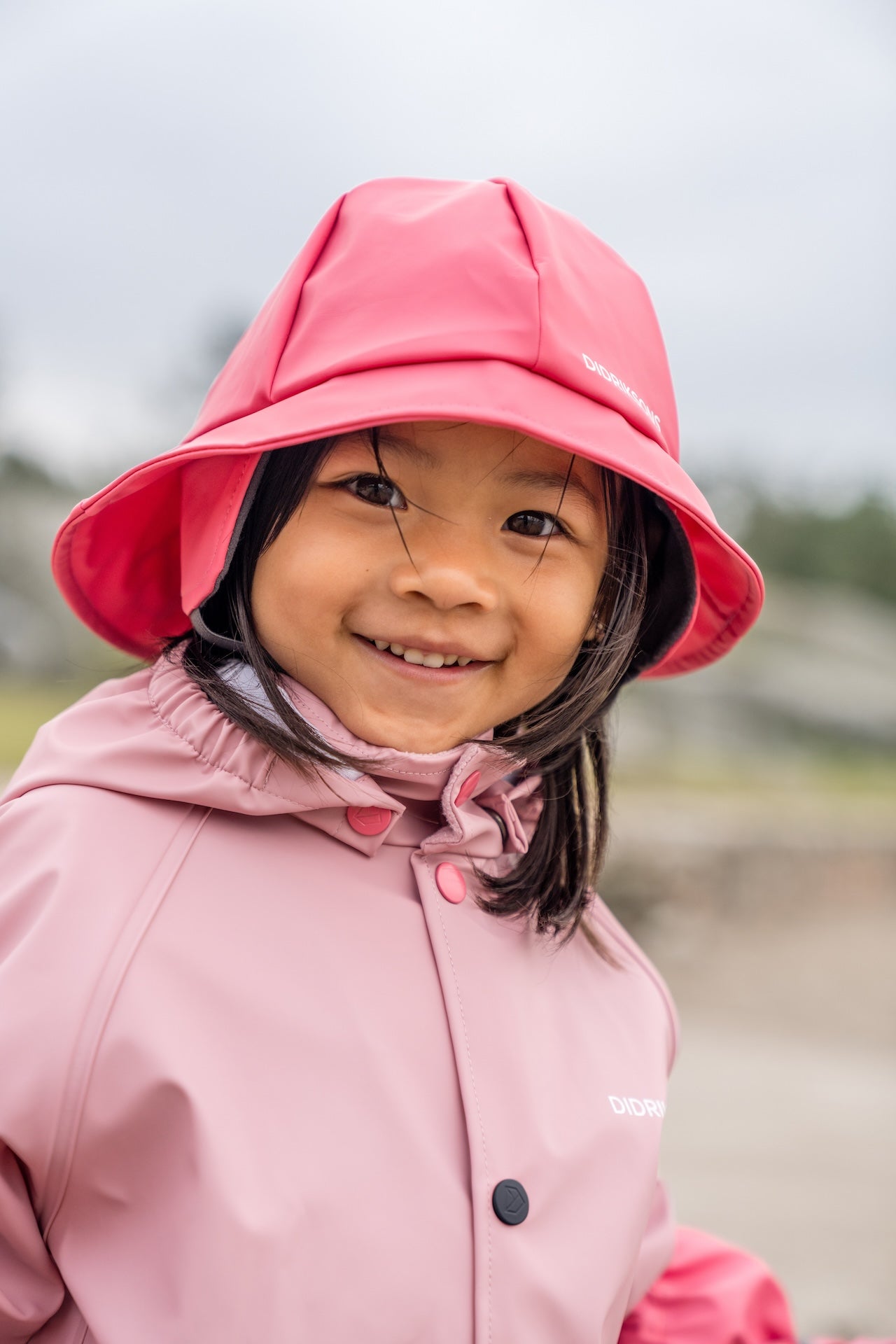 Child wearing a pink raincoat and hat with a blurred outdoor background