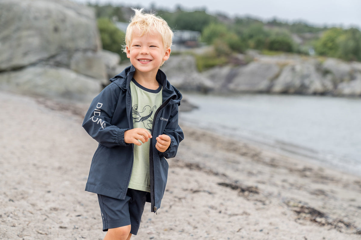 Child miling on a rocky beach, wearing a navy blue G-1000 hooded jacket over a light green graphic t-shirt and matching navy shorts.