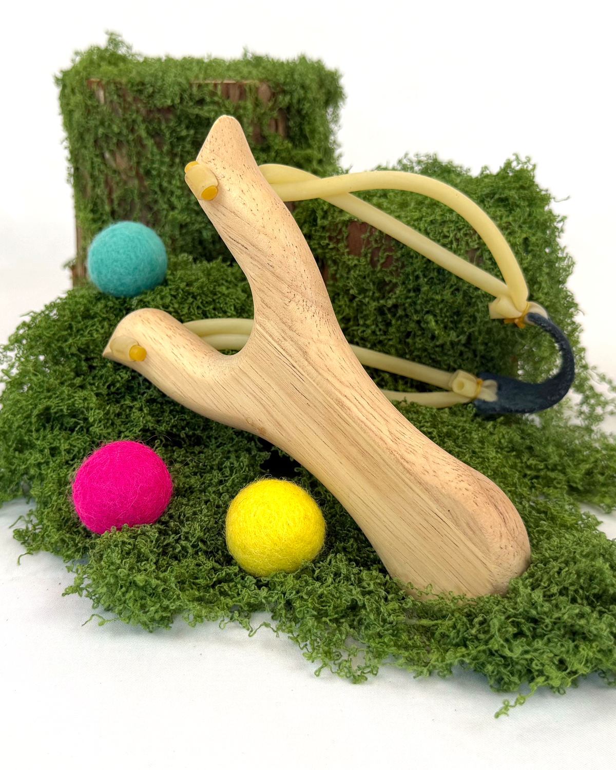 A natural wood Y-shaped slingshot with elastic bands, displayed on a bed of green moss alongside colorful pom-poms and a small tree stump prop against a white background.
