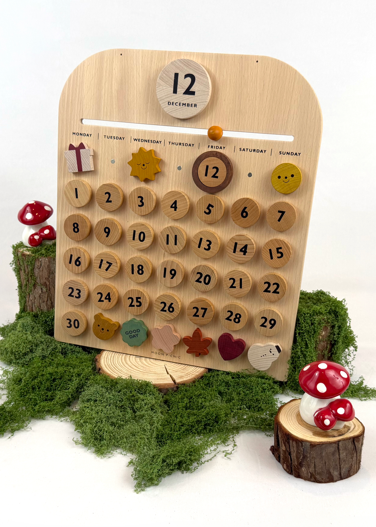A wooden monthly calendar with round numbered pegs and colorful day decorations, displayed on a small wooden stand with green moss and red-and-white mushrooms around it.