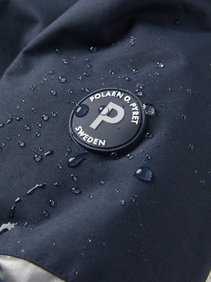 Close-up of a black circular Polarn O. Pivet Sweden logo patch on a dark navy waterproof fabric, with water droplets beaded on the surface showing the inherent waterproof nature of this jacket. 