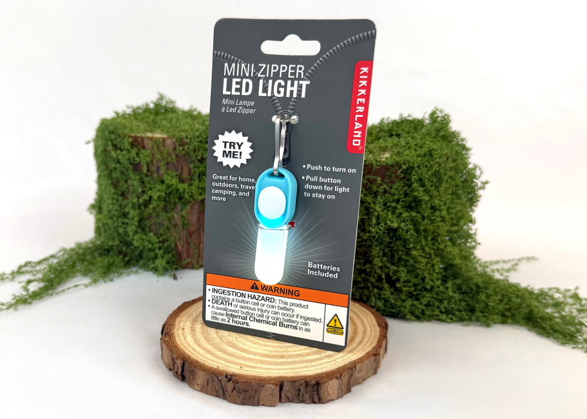 A mini zipper LED light in its retail packaging, displayed on a wooden slice with green moss props against a white background.