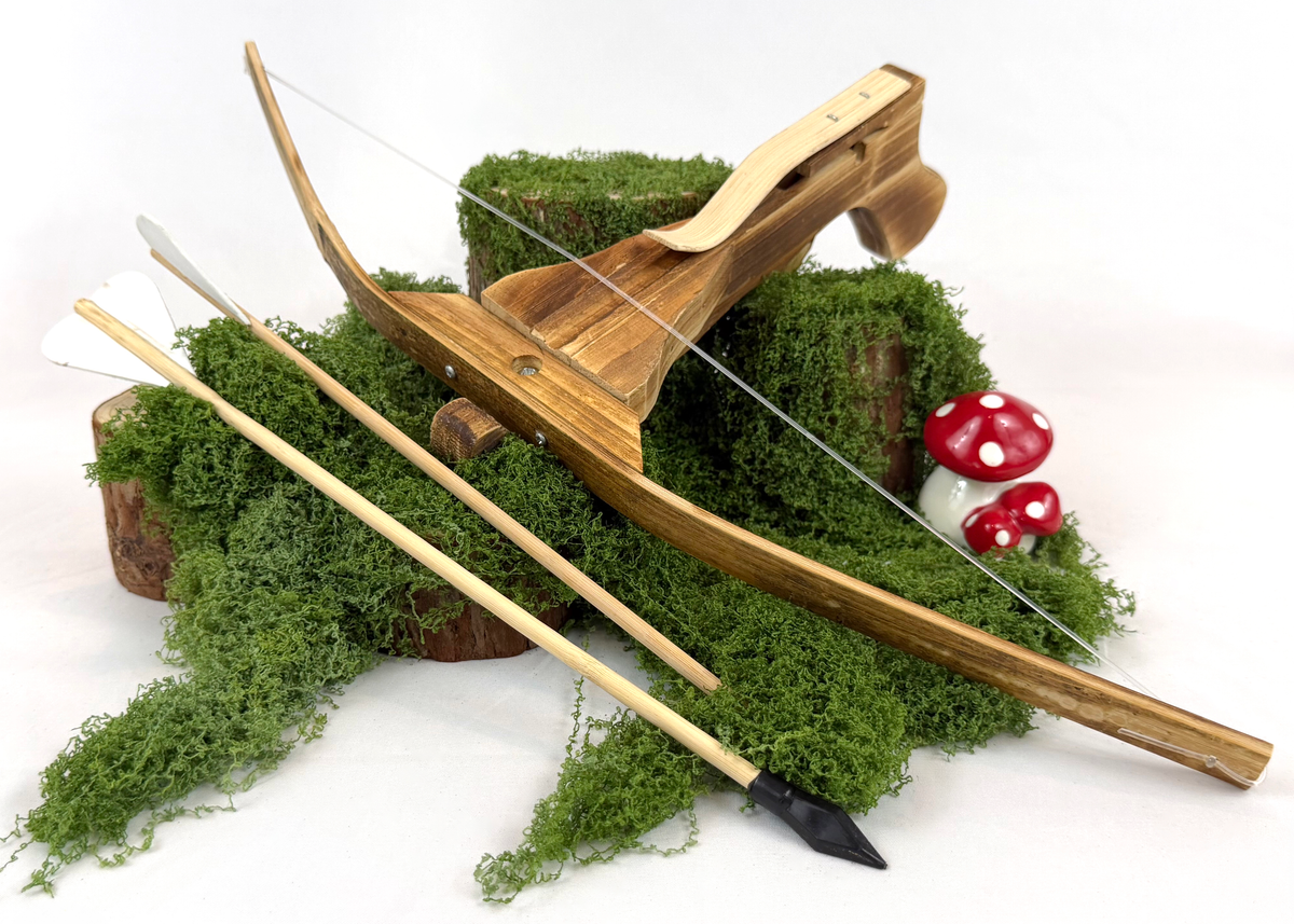 A wooden toy or decorative crossbow with a clear string, displayed on green moss with two arrows and small red-and-white mushrooms on a white background.