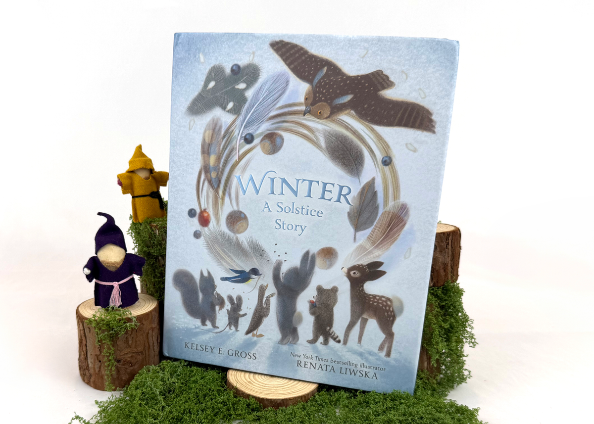 Children's book titled "Winter: A Solstice Story" standing upright on a small wooden stand, surrounded by decorative moss, faux tree stumps, and small figurines against a clean white background.